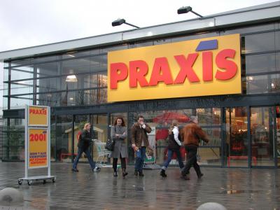 Buitenzijde van een Praxis-winkel met mensen die langs de ingang lopen en een reclamebord op de voorgrond.