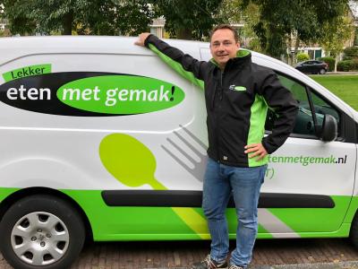 Man in een zwart-groene jas leunt tegen een bestickerde Eten met gemak bestelwagen op straat.