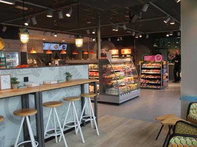 Interieur van een moderne SPAR express winkel met snacks, dranken en tafels met barkrukken in een lichte omgeving.
