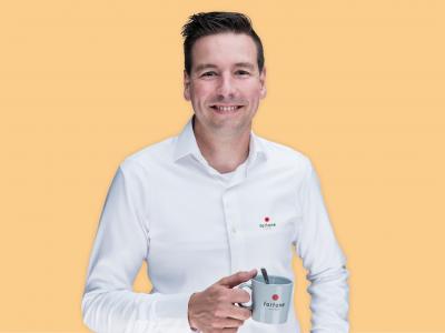 Man in een witte Fortune Coffee-overhemd houdt een grijze mok vast tegen een oranje achtergrond.