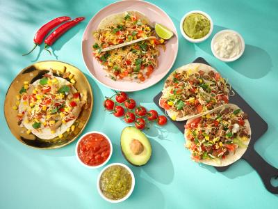 Verschillende kleurrijke taco's met tomaat, kaas, maïs en koriander, geserveerd met sausjes en avocado op een turquoise ac...