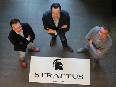 Drie mannen in pakken kijken omhoog naast een Straetus incasso-bord op een grijze tegelvloer.