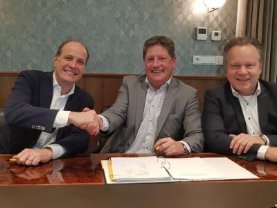 Drie mannen in zakelijke kleding schudden handen rondom een getekend contract, met een Jumbo-logo op de achtergrond.