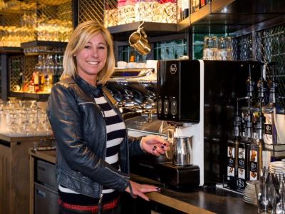 Vrouw in leren jack maakt koffie in een moderne koffiebar met diverse siroopflessen en glazen op de achtergrond.