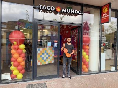 Een man staat voor de ingang van Taco Mundo Amsterdam-Noord met kleurrijke ballonversieringen.