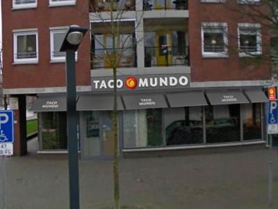 Voorgevel van een Taco Mundo winkel met opvallend logo op straatniveau in een stedelijke omgeving.