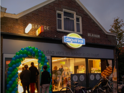Dagwinkel met ballonnenboog bij de ingang en mensen die naar binnen gaan, met fietsen geparkeerd voor de winkel.