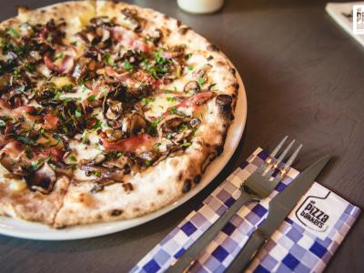 Pizza met paddenstoelen en spek op een bord naast bestek en een servet met De Pizzabakkers-logo.