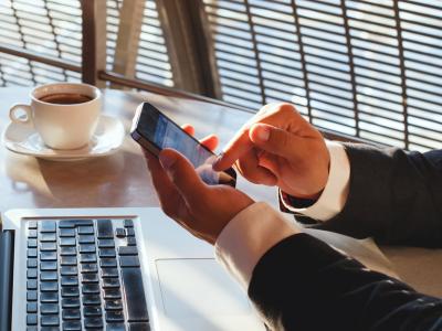 Man in pak gebruikt smartphone in een café terwijl een laptop en kop koffie op tafel staan.