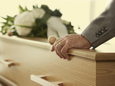 Hand van een man in pak rust op een kist met bloemenkrans op de achtergrond.