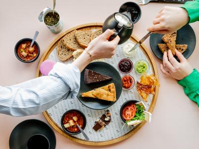 Gezellig gedekte tafel met diverse broodjes, smeersels, taartjes, en handen die thee inschenken en broodjes bereiden.