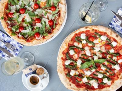 Twee ambachtelijke pizza's met toppings van rucola, tomaat en kaas op een tafel met drankjes en een espresso.