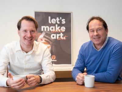 Twee mannen zitten aan een houten tafel met koffiekopjes, met een poster op de achtergrond.