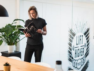Vrouw leest een tijdschrift bij een glazen wand met de tekst Lift Higher en een kantoorplant op de achtergrond.