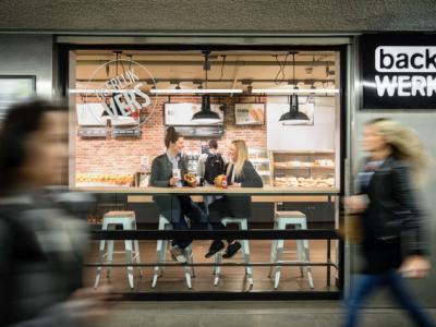 Twee mensen drinken koffie aan een tafel in een backWERK-vestiging, met voorbijgangers op de voorgrond.