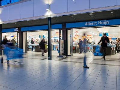 Vooraanzicht van een druk bezochte Albert Heijn-winkel met klanten bij de kassa's en winkelwagens in beweging.