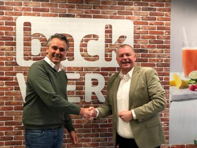 Twee heren schudden handen voor een backWERK-logo op een muur van bakstenen.