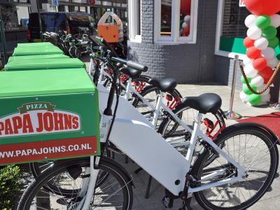 Rij elektrische bezorgfietsen met Papa John’s logo, opgesteld voor winkel met feestelijke ballonnen.