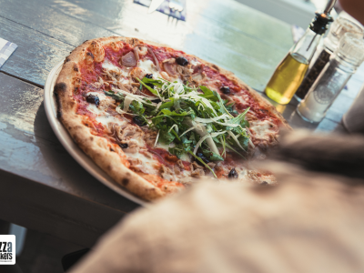 Versierde pizza met rucola op een houten tafel bij De Pizzabakkers, inclusief olijfolie en servet met logo.