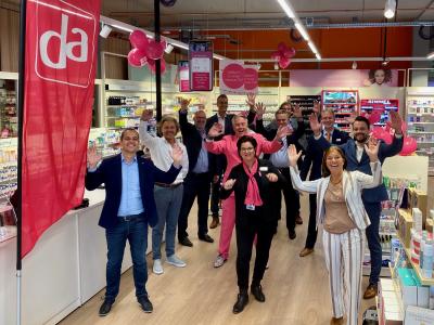 Groep mensen in een DA-winkel viert feestelijke opening met opgeheven handen en glimlachende gezichten.