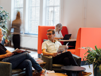 Mensen aan het werk in een moderne kantine met loungestoelen en hoge tafels, gefocust op laptops en overleg.