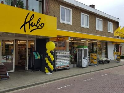 Gevel van een Hubo-winkel met gele en zwarte ballonnen bij de ingang en diverse promotiematerialen buiten.