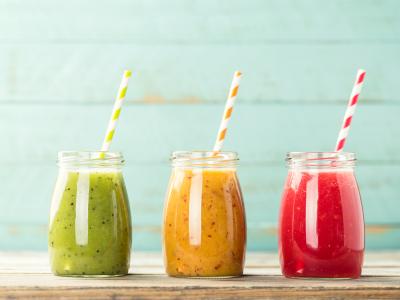 Drie glazen potten met kleurrijke smoothies en rietjes staan op een houten tafel tegen een blauwe achtergrond.