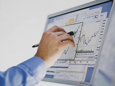 Hand wijst met pen naar oplopende grafiek op computerscherm met meerdere financiële diagrammen.