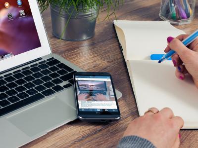 Werkruimte met laptop, smartphone, notitieblok en persoon die notities maakt aan een houten bureau.
