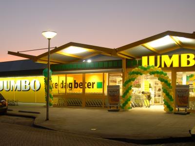 Jumbo supermarkt in avondlicht met verlichte gevel en versierde ingang met groene en gele ballonnen.