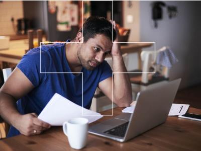 Man in blauwe trui bekijkt bezorgd papieren voor een laptop aan een keukentafel met koffiekopje.