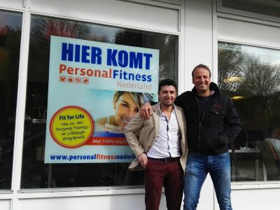 Twee mannen poseren voor een raam met een poster van Personal Fitness Nederland.