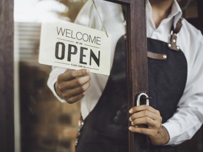 Persoon in schort opent deur met Welcome, We Are Open bordje zichtbaar.