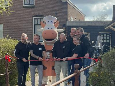 Mensen knippen een rood lint door bij de opening van een nieuwe Pannenkoe-vestiging, met een koe in kostuum op de achtergr...
