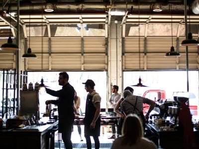 Mensen werken samen in een café met industriële uitstraling en grote ramen, terwijl klanten aan de balie wachten.