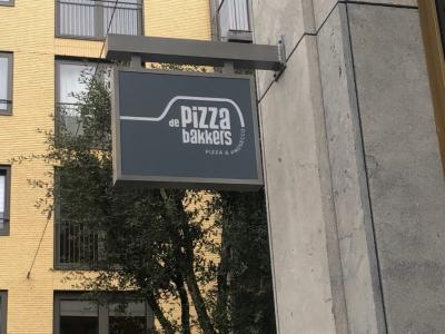 Uithangbord van De Pizzabakkers aan de gevel van een gebouw in een stedelijke omgeving.