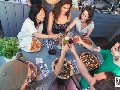 Mensen proosten met glazen rondom een tafel vol pizza's bij De Pizzabakkers in de Twijnstraat.