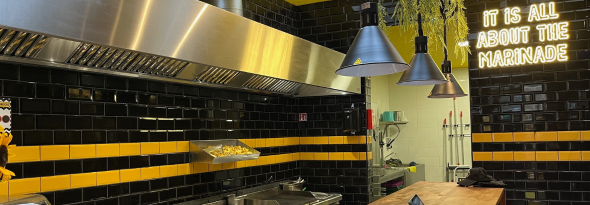 Interieur van een keuken met zwarte en gele tegels, roestvrijstalen afzuigkap en een neonbord met de tekst over marinade.
