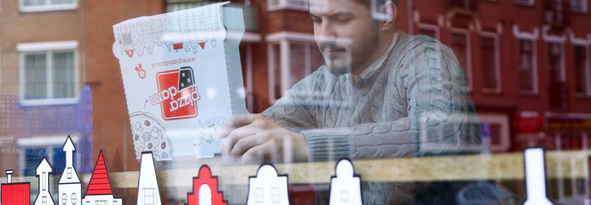 Man binnen bij raam met pizzadoos van Pizzadam, weerspiegeling van appartementen zichtbaar in het glas.