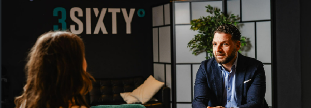 Twee mensen in gesprek aan een tafel in een modern kantoor met de logo's gewoan su. en 3SIXTY op de achtergrond.