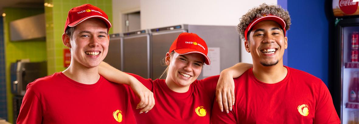 Drie lachende Taco Mundo-medewerkers in rode uniformen in een keukenomgeving.
